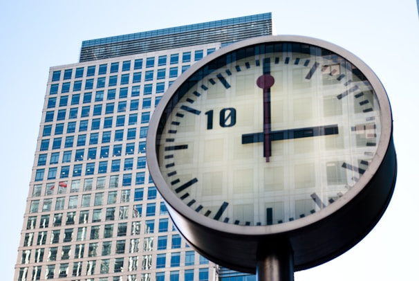 A large, modern clock is prominently displayed in the foreground, with a high-rise office building featuring numerous windows in the background. The clock face is white with black hour and minute markers, and a red second hand.