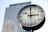A large, modern clock is prominently displayed in the foreground, with a high-rise office building featuring numerous windows in the background. The clock face is white with black hour and minute markers, and a red second hand.