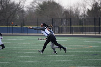 Two athletes are engaged in a competitive lacrosse game on a sports field. One player is in mid-action, extending their arm to catch or throw an orange lacrosse ball. The field is marked with multiple lines, and a fence and trees can be seen in the background.