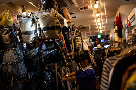 A narrow aisle in a store filled with a variety of backpacks and bags hanging on shelves. A person appears to be shopping or arranging items, surrounded by colorful and patterned bags. The store is brightly lit with overhead lights creating a warm ambiance.