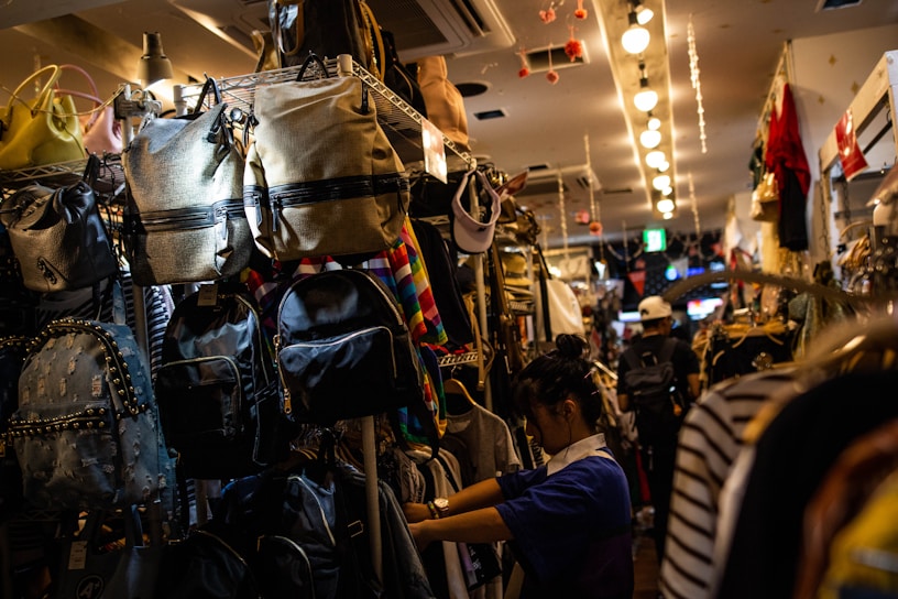 A narrow aisle in a store filled with a variety of backpacks and bags hanging on shelves. A person appears to be shopping or arranging items, surrounded by colorful and patterned bags. The store is brightly lit with overhead lights creating a warm ambiance.