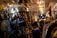 A narrow aisle in a store filled with a variety of backpacks and bags hanging on shelves. A person appears to be shopping or arranging items, surrounded by colorful and patterned bags. The store is brightly lit with overhead lights creating a warm ambiance.