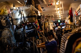 A narrow aisle in a store filled with a variety of backpacks and bags hanging on shelves. A person appears to be shopping or arranging items, surrounded by colorful and patterned bags. The store is brightly lit with overhead lights creating a warm ambiance.