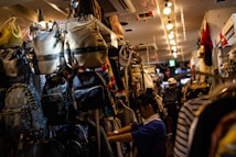 A narrow aisle in a store filled with a variety of backpacks and bags hanging on shelves. A person appears to be shopping or arranging items, surrounded by colorful and patterned bags. The store is brightly lit with overhead lights creating a warm ambiance.