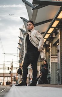 A man in a light trench coat and dark jeans stands confidently on a platform at a train station. A plane is visible in the sky above a series of metal and glass canopies. Other individuals can be seen in the background, some sitting and others standing.