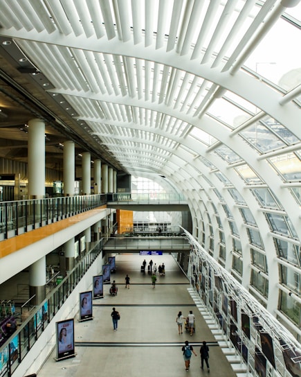 A spacious, modern interior with a high, curved ceiling composed of numerous white slats. The large windows on the right allow ample natural light to filter in. The space features two levels, with glass railings dividing them, and multiple people walking along the ground floor. Large advertising displays line the walls on both levels, and the environment feels airy and open.