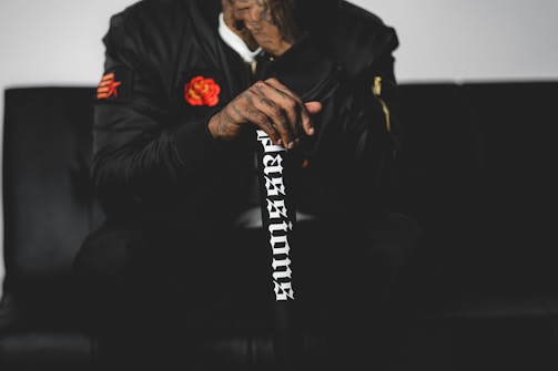 A person wearing a black jacket with a rose patch is seated on a black surface. The individual is holding a black walking stick that has white lettering reading 'passions'. The jacket has distinct patches, including a red flower on the left side.