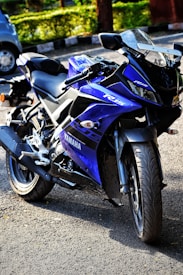 A blue sport motorcycle with a sleek and aerodynamic design is parked on a street. The motorcycle features prominent branding and detailing with clear lines and visible mechanical components. It is positioned at an angle, highlighting its large front tire and handlebars. The background shows a blurred car and greenery, adding contrast to the scene.