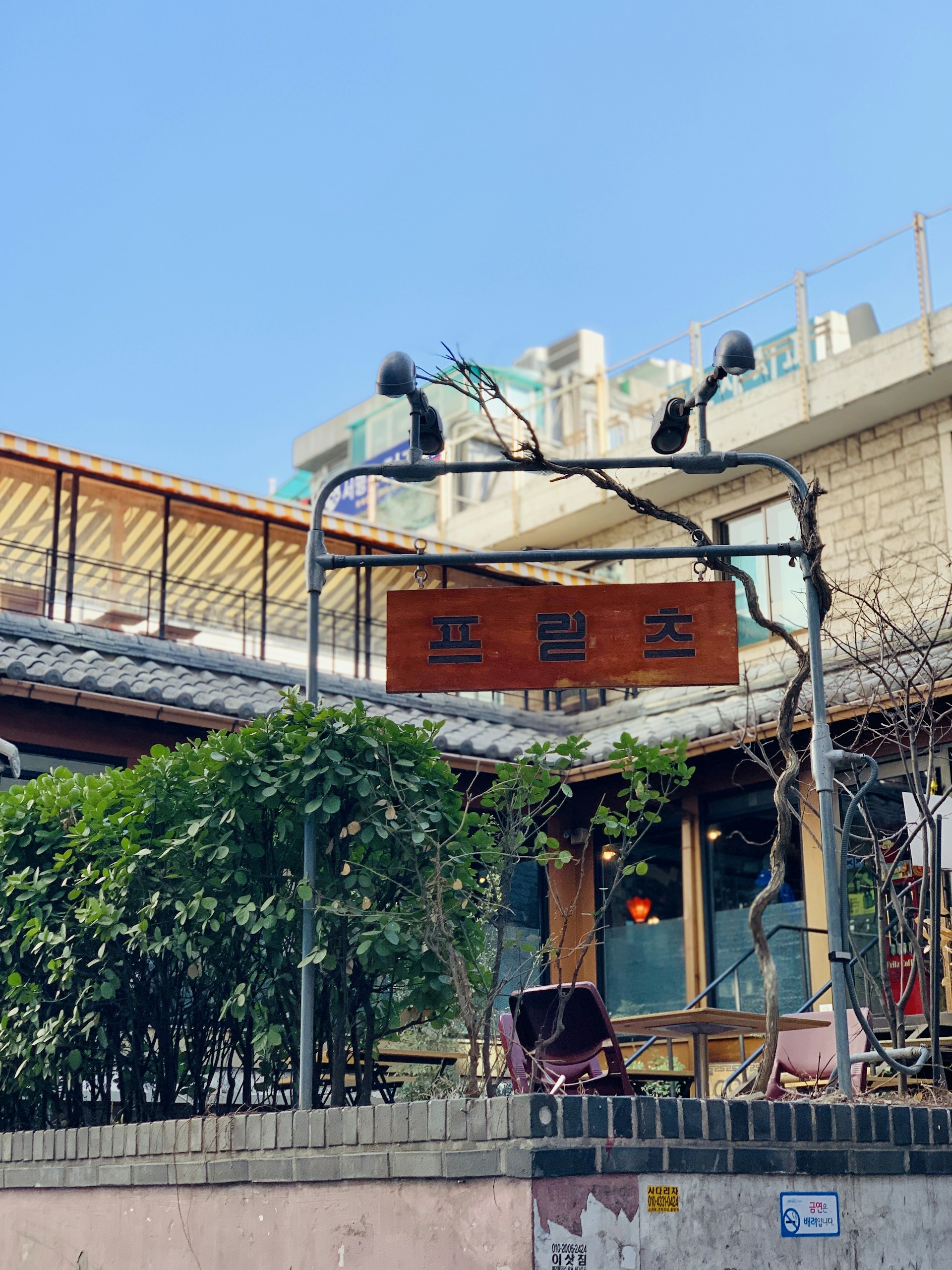 A wooden sign with a word in Korean hangs from a metal frame with intertwining vines. Under the sign, there are green bushes and a glimpse of a patio area with plants and what appears to be tables and chairs. In the background, there are multiple buildings with a terrace and other architectural features. The sky is clear and blue.