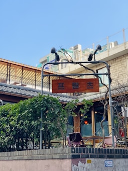 A wooden sign with a word in Korean hangs from a metal frame with intertwining vines. Under the sign, there are green bushes and a glimpse of a patio area with plants and what appears to be tables and chairs. In the background, there are multiple buildings with a terrace and other architectural features. The sky is clear and blue.