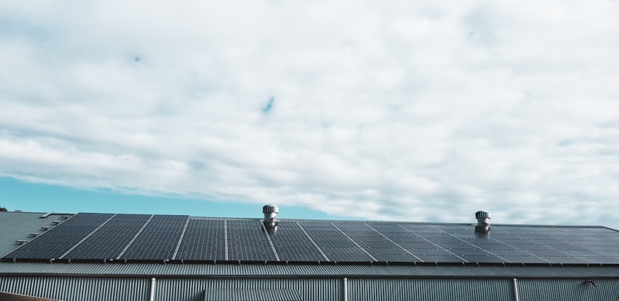 A rooftop covered with a large array of solar panels beneath a cloudy sky. The roof features two metallic vent structures protruding from the panels. The scene is outdoors and the weather appears overcast.