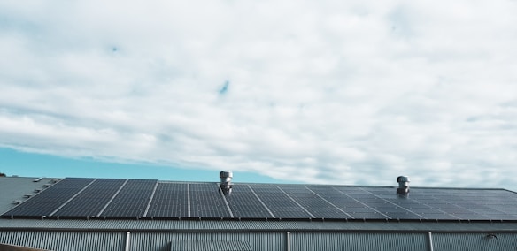 A rooftop covered with a large array of solar panels beneath a cloudy sky. The roof features two metallic vent structures protruding from the panels. The scene is outdoors and the weather appears overcast.