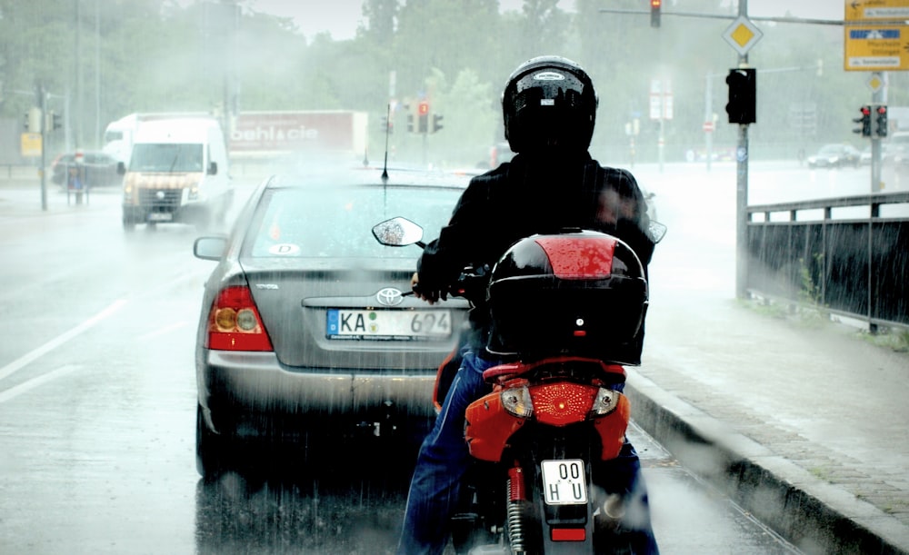 man riding red motor scooter with gray Toyota sedan in front of him during rainy daytime