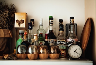 A collection of various liquor bottles is arranged on a white shelf, including recognizable brands. To the left, there is a paper bag with dried herbs and a golden box with a grape design. In the foreground, there are glass cups with wooden bases. An antique-style alarm clock is placed to the right, next to a wooden tray leaning against the wall.