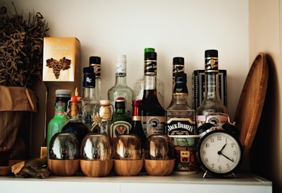 A collection of various liquor bottles is arranged on a white shelf, including recognizable brands. To the left, there is a paper bag with dried herbs and a golden box with a grape design. In the foreground, there are glass cups with wooden bases. An antique-style alarm clock is placed to the right, next to a wooden tray leaning against the wall.