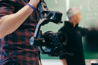 A person holds a professional camera mounted on a stabilizing gimbal. The person is wearing a checkered shirt and a blue wristband. In the blurred background, another individual appears to be writing on a chalkboard.