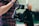 A person holds a professional camera mounted on a stabilizing gimbal. The person is wearing a checkered shirt and a blue wristband. In the blurred background, another individual appears to be writing on a chalkboard.