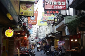 A bustling street scene in an Asian city with numerous colorful signs and storefronts. People are walking and riding scooters, while shops display various goods. The atmosphere is lively and crowded, suggesting a busy commercial area.