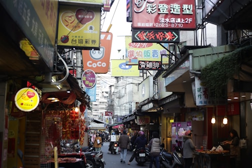 A bustling street scene in an Asian city with numerous colorful signs and storefronts. People are walking and riding scooters, while shops display various goods. The atmosphere is lively and crowded, suggesting a busy commercial area.