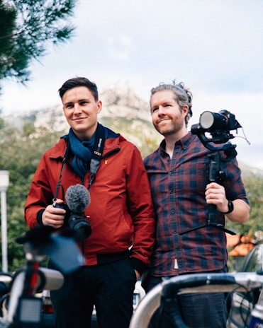 Two individuals are standing outdoors, each holding video or photography equipment. One person is dressed in a red jacket and scarf, holding a camera with a microphone attached. The other person is wearing a plaid shirt and is holding a camera on a stabilizer. In the background, there is greenery and a mountain range. The scene appears to be casual, possibly during a filming session or photography outing.