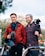Two individuals are standing outdoors, each holding video or photography equipment. One person is dressed in a red jacket and scarf, holding a camera with a microphone attached. The other person is wearing a plaid shirt and is holding a camera on a stabilizer. In the background, there is greenery and a mountain range. The scene appears to be casual, possibly during a filming session or photography outing.