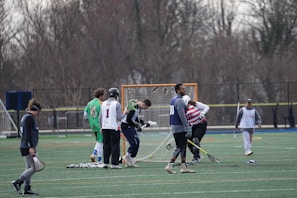 A group of lacrosse players are gathered on an outdoor field with lacrosse gear and sticks. Some players are dressed in jerseys, with helmets and gloves, standing around a lacrosse goal net. The field is surrounded by trees without leaves, indicating a possible early spring or late autumn setting. One player on the left is walking away, while another in a green jersey appears to talk to someone. The goal net has some equipment laying nearby.