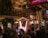 A crowded festive street scene with people gathered around a decorated area featuring a booth labeled 'Santa's Workshop'. Lights and ornaments are visible on a nearby tree, with a balcony overlooking the crowd where more people are watching. Nearby businesses include a restaurant and greenery adds to the festive atmosphere.