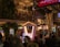 A crowded festive street scene with people gathered around a decorated area featuring a booth labeled 'Santa's Workshop'. Lights and ornaments are visible on a nearby tree, with a balcony overlooking the crowd where more people are watching. Nearby businesses include a restaurant and greenery adds to the festive atmosphere.