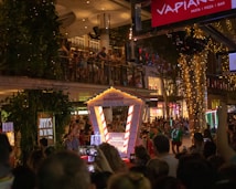 A crowded festive street scene with people gathered around a decorated area featuring a booth labeled 'Santa's Workshop'. Lights and ornaments are visible on a nearby tree, with a balcony overlooking the crowd where more people are watching. Nearby businesses include a restaurant and greenery adds to the festive atmosphere.