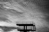 A silhouette of a diving platform stands against a dramatic sky filled with textured and wavy clouds.