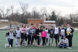 A group of people is standing on a sports field holding lacrosse sticks. They appear to be a lacrosse team, dressed in various athletic outfits, with some wearing jerseys. The setting is outdoors, with a few trees and a building visible in the background. The sky is overcast.