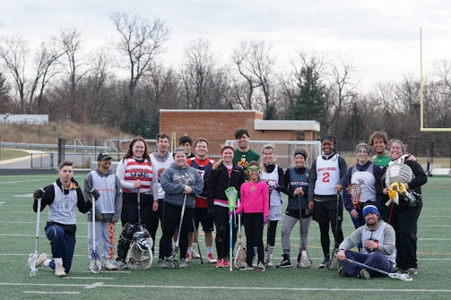 A group of people is standing on a sports field holding lacrosse sticks. They appear to be a lacrosse team, dressed in various athletic outfits, with some wearing jerseys. The setting is outdoors, with a few trees and a building visible in the background. The sky is overcast.