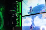 Two gaming monitors display an action scene from a video game featuring a character with purple hair in a lush outdoor setting. The gaming PCs are mounted on the wall with glowing green LED lights, and the room is dimly lit to emphasize the gaming setup.