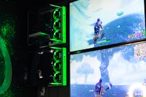 Two gaming monitors display an action scene from a video game featuring a character with purple hair in a lush outdoor setting. The gaming PCs are mounted on the wall with glowing green LED lights, and the room is dimly lit to emphasize the gaming setup.
