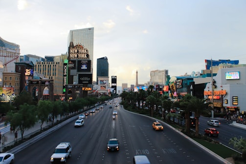 A bustling urban scene featuring a wide boulevard with multiple cars driving in both directions. The sides of the street are lined with well-known hotels and casinos, each displaying brightly-lit signs and advertisements. Among the notable structures is a replica of the Eiffel Tower. The atmosphere is lively and vibrant, with abundant palm trees adding a touch of greenery.