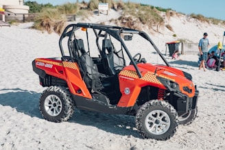 A bright orange beach rescue vehicle is parked on the sand, featuring distinct checkered patterns and branding. In the background, a beach scene unfolds with a few people, including a man standing nearby and others sitting or playing in the sand. Surrounding the vehicle, sand dunes and some grassy areas are visible.
