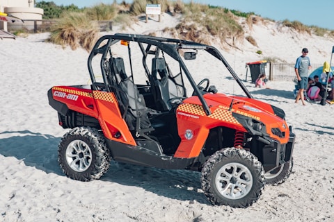 A bright orange beach rescue vehicle is parked on the sand, featuring distinct checkered patterns and branding. In the background, a beach scene unfolds with a few people, including a man standing nearby and others sitting or playing in the sand. Surrounding the vehicle, sand dunes and some grassy areas are visible.