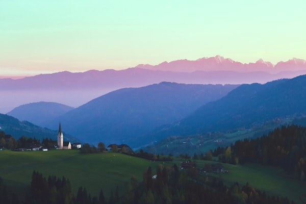 A serene landscape featuring rolling hills bathed in soft light. A church with a tall spire is nestled among the greenery in the foreground. The horizon is layered with majestic mountains, slightly obscured by mist, under a pastel-colored sky.