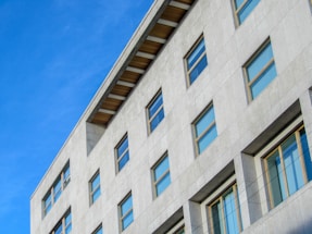 A modern multi-story building with a light gray facade and numerous rectangular windows. The structure features a flat roof with exposed beams and is set against a clear blue sky, giving it a clean and minimalistic appearance.