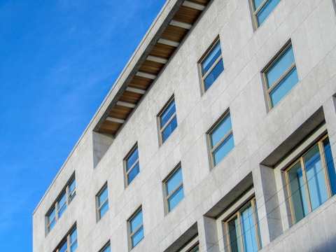 A modern multi-story building with a light gray facade and numerous rectangular windows. The structure features a flat roof with exposed beams and is set against a clear blue sky, giving it a clean and minimalistic appearance.