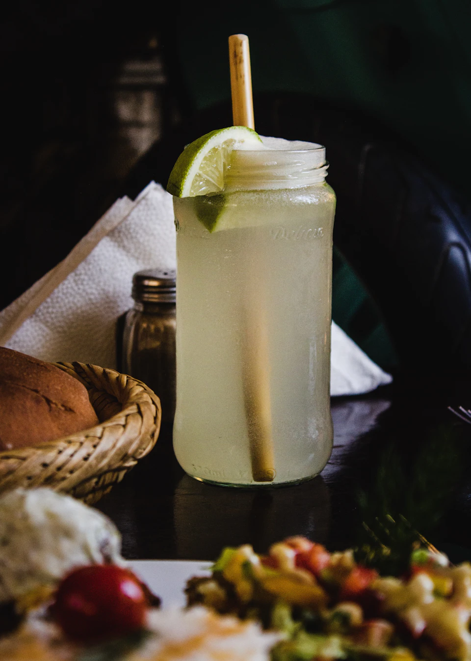 The image shows a tall glass jar filled with a pale yellow liquid, likely lemonade or tea, placed on a dark wooden table. The jar has a straw protruding from it and is topped with a slice of lime. To the left of the jar, there's a basket containing bread and other food items. On the right side of the jar, there are two small bowls filled with different types of food.