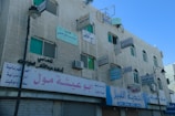 An old, multi-story concrete building features numerous advertising signs in Arabic, with some signs mounted perpendicular to the walls and others flat against them. The building has several windows with green-tinted glass, and air conditioning units are visible under some windows. The ground floor hosts shops with closed roller shutters, and an ornate street lamp is visible in front of the building. The sky is clear and blue, providing a contrasting backdrop to the building.