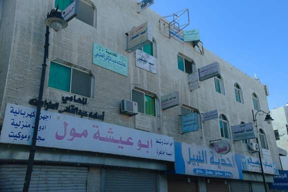 An old, multi-story concrete building features numerous advertising signs in Arabic, with some signs mounted perpendicular to the walls and others flat against them. The building has several windows with green-tinted glass, and air conditioning units are visible under some windows. The ground floor hosts shops with closed roller shutters, and an ornate street lamp is visible in front of the building. The sky is clear and blue, providing a contrasting backdrop to the building.