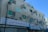An old, multi-story concrete building features numerous advertising signs in Arabic, with some signs mounted perpendicular to the walls and others flat against them. The building has several windows with green-tinted glass, and air conditioning units are visible under some windows. The ground floor hosts shops with closed roller shutters, and an ornate street lamp is visible in front of the building. The sky is clear and blue, providing a contrasting backdrop to the building.