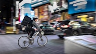 A cyclist wearing a helmet is riding through a bustling city street at night, carrying a large backpack. Bright neon signs and billboards illuminate the surroundings, with blurred motion capturing the fast-paced environment. Cars pass by, contributing to the energetic urban scene.