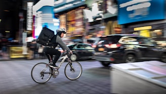 A cyclist wearing a helmet is riding through a bustling city street at night, carrying a large backpack. Bright neon signs and billboards illuminate the surroundings, with blurred motion capturing the fast-paced environment. Cars pass by, contributing to the energetic urban scene.