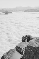 A stunning landscape features rugged, snow-capped mountains in the foreground and a vast sea of clouds extending into the distance. The horizon is lined with a range of distant peaks, creating an impressive sense of scale and depth.