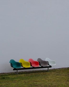 A row of five colorful chairs attached to a metal frame is placed against a plain white wall. The chairs are in distinct colors: teal, yellow, red, gray, and white. The setting appears to be outdoors, with a patch of grassy ground extending across the bottom of the image.