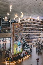 An indoor library with tall, curved bookshelves filled with books. The shelves extend to almost the ceiling, and there's a colorful mural on one side. Star-shaped lights hang from the ceiling. People are seated on benches and walking around, adding a vibrant atmosphere.
