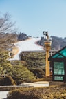 A snow-covered ski slope with numerous people skiing down, surrounded by evergreen trees. In the foreground, a whimsical structure resembling a pirate-themed lookout tower with a skull and crossbones flag is visible. A modern glass gazebo is positioned to the right. The background features a clear blue sky and distant tree-covered hills.